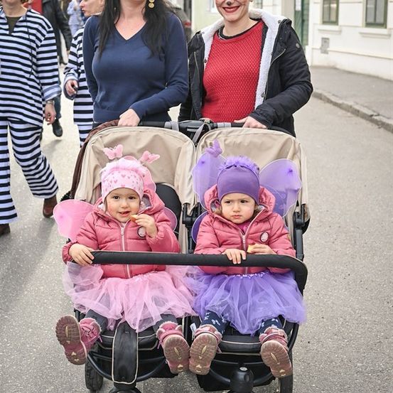 Zwei Frauen schieben einen Doppelkinderwagen mit zwei Kleinkindern in Feenkostümen. Sie lächeln und scheinen einen Umzug zu genießen. Hinter ihnen laufen Menschen in Kostümen die Straße entlang.