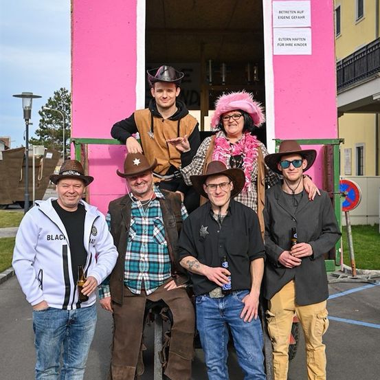 Eine Gruppe von Menschen in Cowboyhüten und Kostümen posiert vor einem rosa Gebäude mit einem Schild, auf dem 'LANGE' steht.