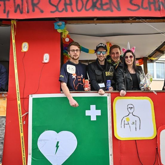 Eine Gruppe von Menschen steht in einem Stand mit rotem Hintergrund und Dekorationen. Sie lächeln und posieren für ein Foto. Der Stand hat ein grünes Banner mit einem Herz und einem Blitz. Der Mann in der Mitte trägt ein SWAT-Shirt. Der Stand hat ein gelbes Warnband und ein Schild, das 'AF, WIR SCHAUEN SCHA' besagt.