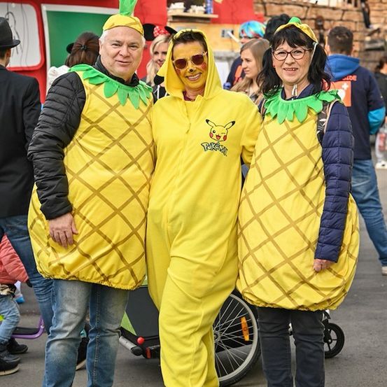 Drei Menschen in gelben Ananas-Kostümen stehen in einem öffentlichen Bereich, mit einem Kinderwagen und einer Menschenmenge im Hintergrund.