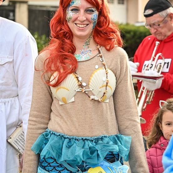 Eine Frau, verkleidet als Arielle aus Die kleine Meerjungfrau, mit roten Haaren und blauem Gesicht, steht vor einem Gebäude, während ein Mann eine Trompete spielt.