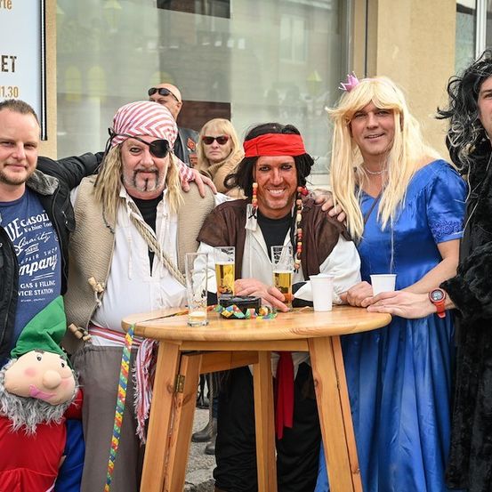 Eine Gruppe von Menschen in verschiedenen Kostümen, darunter ein Pirat, ein Kobold und eine Frau in einem blauen Kleid, posiert für ein Foto vor einem Holztisch mit Getränken.