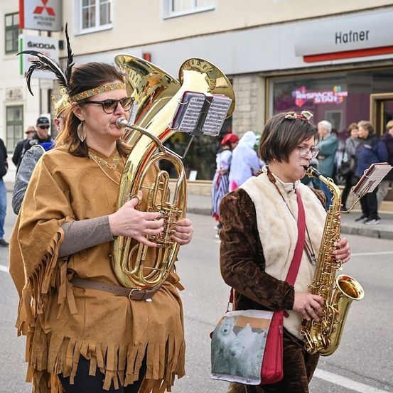 Zwei Frauen in Kostümen spielen Blechblas- und Saxophoninstrumente bei einem Umzug. Die Frau auf der linken Seite trägt eine Federhaube und ein Kleid mit Fransen. Die Frau auf der rechten Seite trägt eine Pelzweste und trägt eine Tasche mit einem Eisbärendesign.