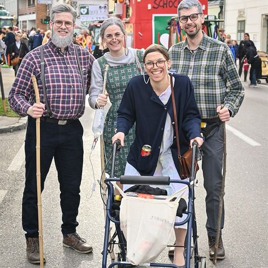 Vier Menschen in Kostümen stehen auf einer Straße. Die Frau in der Mitte schiebt einen Rollator mit einer Tasche. Die beiden Männer halten Stöcke. Im Hintergrund sind eine Menschenmenge und Gebäude zu sehen.