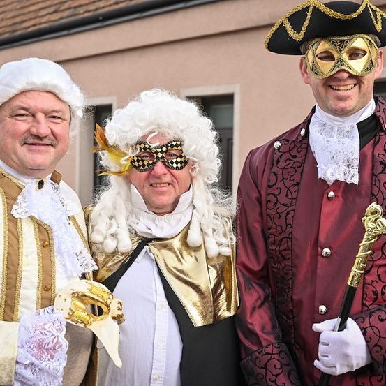 Drei Männer in aufwendigen Kostümen, möglicherweise für ein Fest, stehen lächelnd eng beieinander. Der Mann links trägt eine weiße Perücke und ein weißes und goldenes Kostüm. Der Mann in der Mitte trägt eine weiße Perücke, eine goldene und schwarze Maske und ein goldenes und schwarzes Kostüm. Der Mann rechts trägt einen schwarzen Hut, ein rotes und goldenes Kostüm und hält einen goldbesetzten Stock.