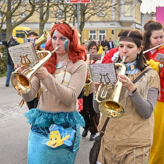 Eine Gruppe von Menschen in Kostümen spielt auf der Straße Musikinstrumente. Eine Frau ist als Arielle aus Die kleine Meerjungfrau verkleidet.