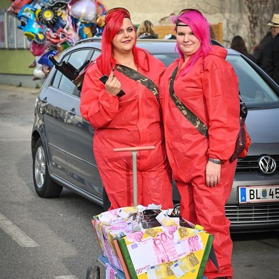 Zwei Frauen in roten Overalls stehen neben einem Auto, eine schiebt einen mit Geld gefüllten Schubkarren. Sie haben pinkfarbene Haare und lächeln. Ein Auto mit dem Kennzeichen 'BL 45' steht hinter ihnen.