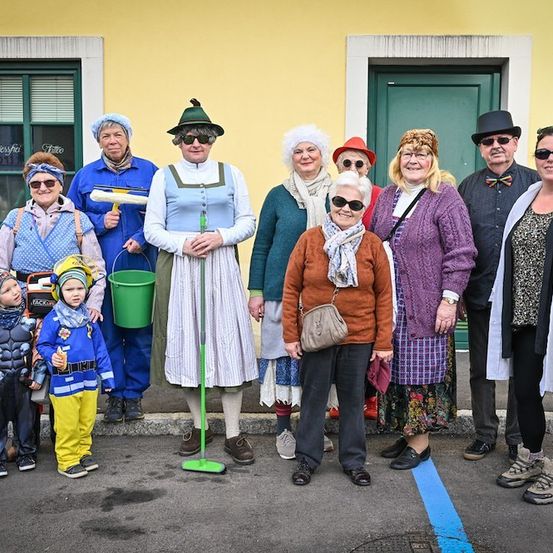 Eine Gruppe von Erwachsenen und Kindern in Kostümen posiert für ein Foto vor einem gelben Gebäude. Die Erwachsenen tragen Hüte, Brillen und verschiedene Accessoires. Die Kinder sind als Thor und Feuerwehrmann verkleidet.