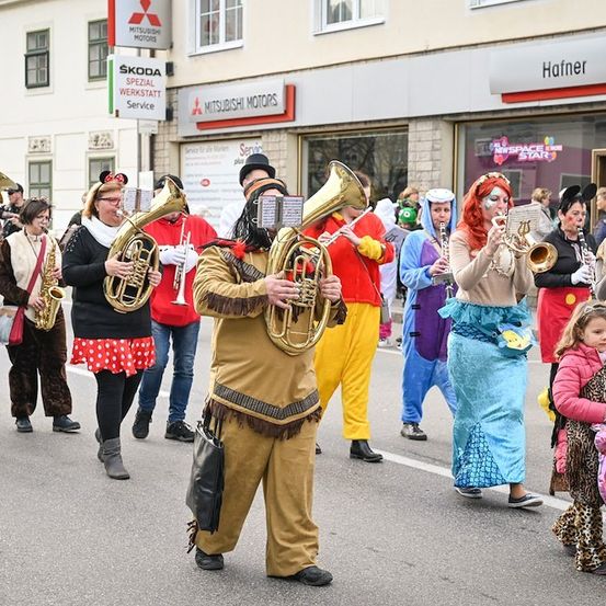 Eine Gruppe von Menschen in Kostümen, die bei einem Umzug Musikinstrumente spielen. Die Kostüme umfassen Minnie Maus, einen Piraten und einen Indianer. Die Instrumente umfassen Trompeten, Saxophone und Tuben. Im Hintergrund befindet sich ein Gebäude mit Schildern für Skoda und Mitsubishi.