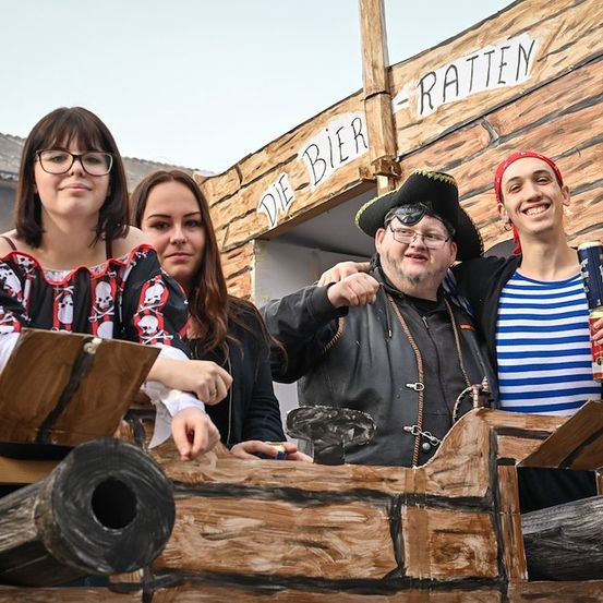 Vier Menschen in Kostümen posieren vor einem hölzernen Piratenschiff. Der Mann in der Mitte trägt einen Piratenhut und hält eine Dose. Die Frauen auf der linken Seite tragen eine Brille und ein Kleid mit Totenkopf-Motiv. Der Mann auf der rechten Seite trägt ein gestreiftes Hemd und ein rotes Kopftuch.