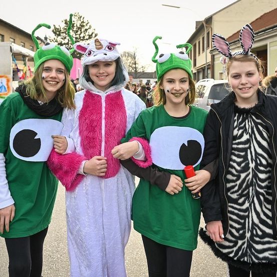 Vier Mädchen in Kostümen posieren für ein Foto auf einer Straße mit Gebäuden und einer Menschenmenge im Hintergrund.