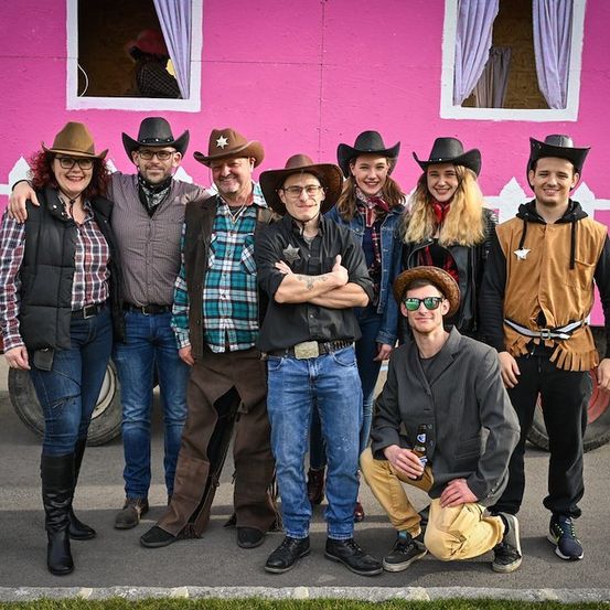 Eine Gruppe von Menschen in Cowboy-Kleidung posiert vor einem rosa Haus. Einige stehen, andere sitzen auf dem Boden.