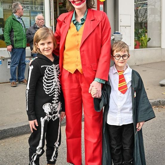 Eine Person, die als Joker verkleidet ist, steht zwischen zwei Kindern, eines als Skelett und das andere als Zauberer gekleidet. Sie sind auf einer Straße mit einem Geschäft im Hintergrund.