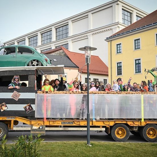 Ein bunter Umzugswagen mit einem grünen Auto auf dem Dach, der eine Menschenmenge in farbenfrohen Kostümen transportiert, fährt eine Straße hinunter, mit Gebäuden im Hintergrund.