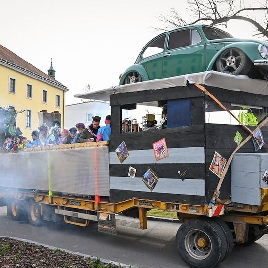 Ein grüner Vintage-Volkswagen Käfer ist auf einem gelben Lastwagen montiert, mit mehreren Personen im Inneren. Der Lastwagen ist mit Aufklebern und Postern verziert. Er fährt auf einer Straße mit einem Gebäude im Hintergrund.