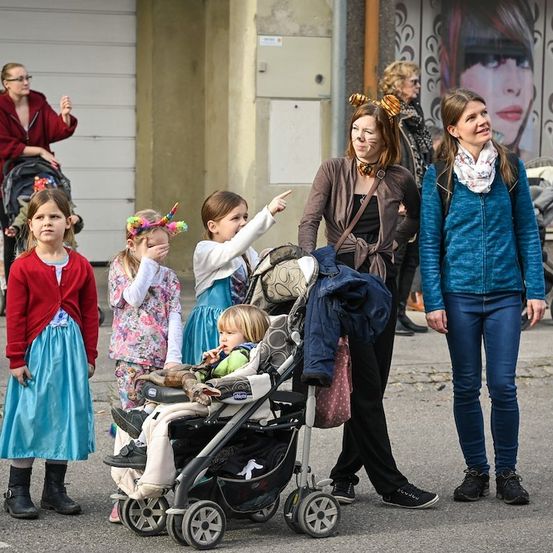 Eine Gruppe von Kindern und Erwachsenen in Kostümen steht auf der Straße. Ein Kind ist in einem Kinderwagen und ein anderes zeigt. Zwei Frauen stehen in der Nähe.