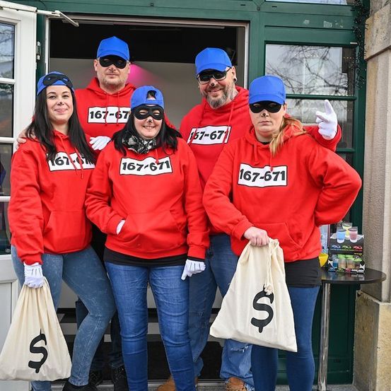 Fünf Personen in roten Kapuzenpullovern mit Nummern und blauen Mützen posieren für ein Foto vor einem Gebäude. Sie halten weiße Beutel mit Dollarzeichen.