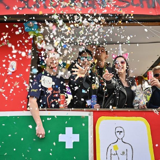 Eine Gruppe von Menschen in einem dekorierten Stand wirft Konfetti, wobei eine Person ein Getränk in der Hand hat. Sie lächeln und scheinen zu feiern.