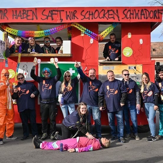 Eine Gruppe von Menschen in passenden blauen Shirts mit einem roten und grünen Wagen dahinter. Der Wagen hat ein Schild, das 'Wir haben Saft, wir schocken Schwadorf' besagt. Die Leute posieren für ein Foto.