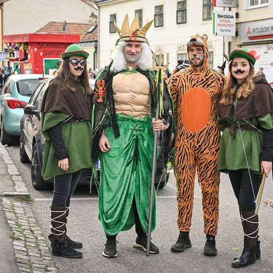 Vier Menschen in Kostümen stehen auf einer Straße während eines Umzugs. Der Mann in der Mitte trägt eine Krone und hält einen Dreizack. Die anderen tragen grüne Umhänge und Masken. Hinter ihnen sind Autos und ein Gebäude mit einem Schild zu sehen.