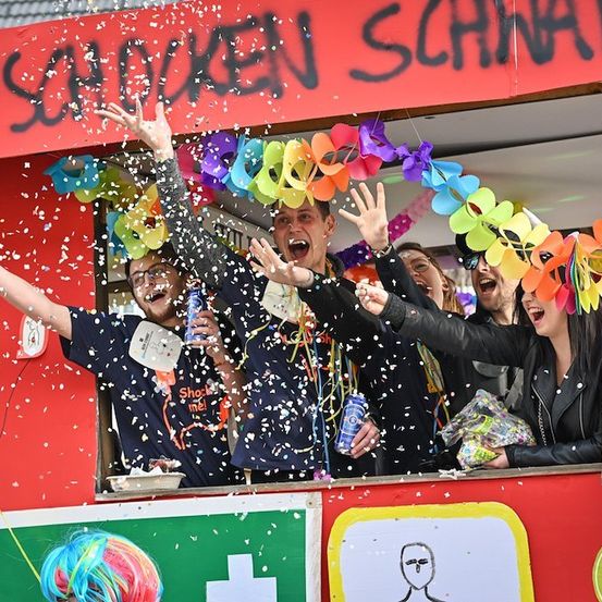 Eine Gruppe von Menschen in einem dekorierten Stand wirft Konfetti und hält Getränke, wobei eine Person eine bunte Perücke trägt.