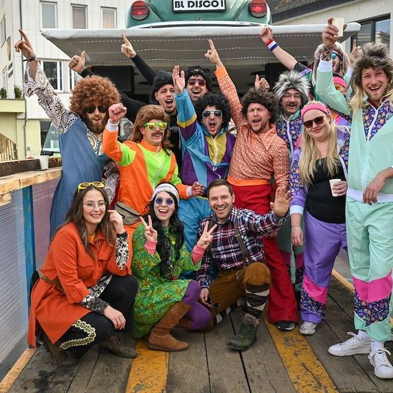 Eine Gruppe von Menschen in farbenfrohen, retro-inspirierten Kostümen posiert für ein Foto auf einer Holzplattform vor einem Bus.