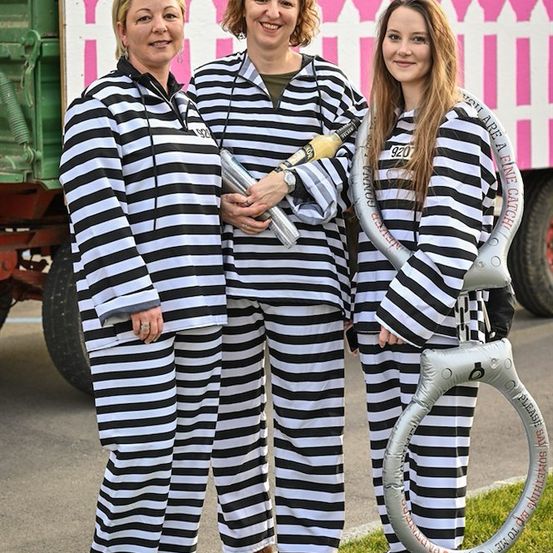 Drei Frauen in schwarz-weiß gestreiften Jumpsuits, lächelnd und posierend für ein Foto mit einem rosafarbenen Lastwagen im Hintergrund.