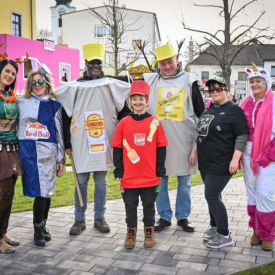 Eine Gruppe von Erwachsenen und einem Kind in Kostümen, wahrscheinlich für ein Festival, stehen auf einem Ziegelweg. Die Kostüme umfassen einen roten Bullen, eine Schweppes-Tonic-Water-Flasche und ein Einhorn.
