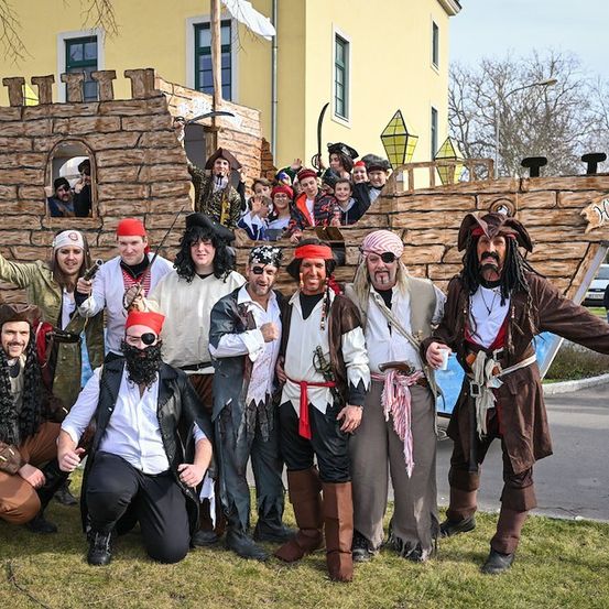 Eine Gruppe von Menschen, die als Piraten verkleidet sind, posiert vor einem Piratenschiffsfloat, wobei einige auf dem Schiff und andere auf dem Gras stehen.