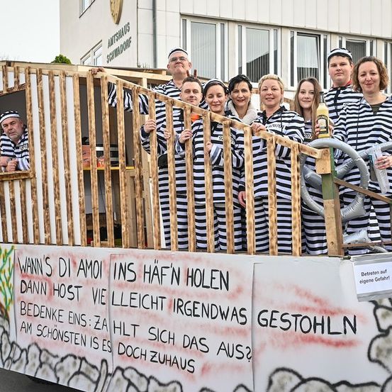 Eine Gruppe von Menschen in schwarz-weiß gestreiften Gefängnisuniformen steht auf einem Wagen, einer hält eine Flasche und ein anderer ein Lenkrad. Dahinter steht ein Holzzaun und ein Gebäude mit einem Schild.