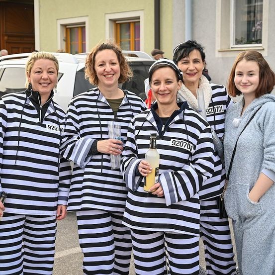 Fünf Frauen in schwarzen und weißen gestreiften Gefängnisoveralls, die lächelnd für ein Foto posieren. Eine Frau hält eine Flasche, und sie stehen vor einem Gebäude.