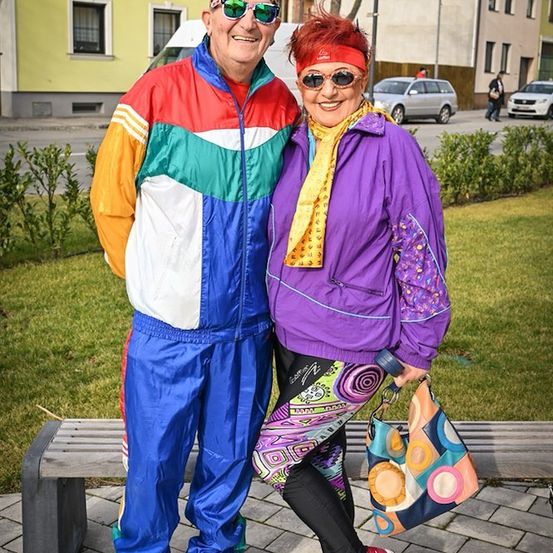 Ein Paar in leuchtender, retro-stilvoller Sportbekleidung steht auf einem gepflasterten Bürgersteig und lächelt für ein Foto.