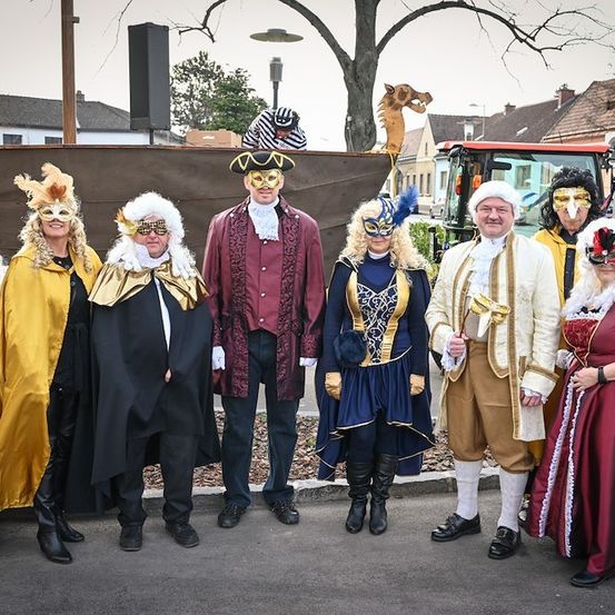 Eine Gruppe von Erwachsenen, die in aufwendigen Kostümen und Masken gekleidet sind, posiert für ein Foto in einer Außenumgebung mit einem Hintergrund aus einem Drachen und Gebäuden.