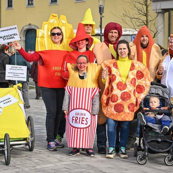 Eine Gruppe von Menschen, die in Essenskostümen wie Pommes Frites, Pizza und Hot Dogs gekleidet sind, steht draußen. Ein Baby in einem Kinderwagen ist ebenfalls anwesend.