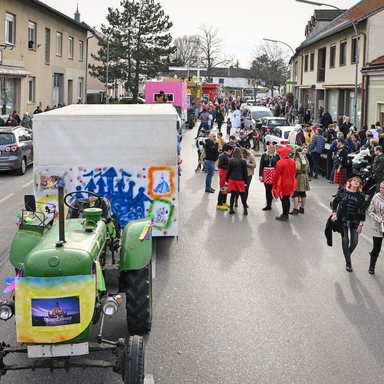 Ein dekorierter Traktor führt eine Parade durch eine Stadtstraße, mit einer Menge Menschen, die entlang der Straße gehen und stehen. Gebäude säumen die Straße, und einige Menschen tragen Kostüme.