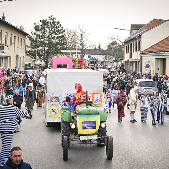 Eine große Menschenmenge in Kostümen und Masken versammelt sich auf einer Straße und beobachtet einen Umzug mit einem geschmückten Traktor an der Spitze. Gebäude und geparkte Autos säumen die Straße.
