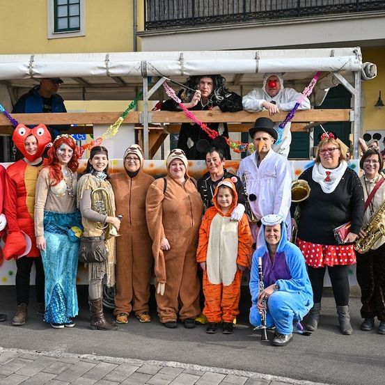 Eine Gruppe von Menschen in verschiedenen Kostümen, einige mit Musikinstrumenten, stehen vor einem geschmückten Wagen, auf dem einige Personen sitzen.
