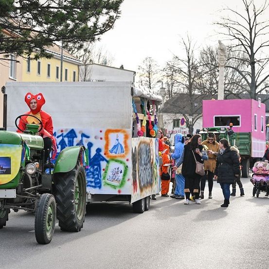 Ein Umzug mit einem grünen Traktor, der einen Anhänger mit bunten Graffiti zieht. Eine Person in einem roten Kostüm fährt den Traktor. Menschen in Kostümen und andere gehen auf der Straße. Gebäude und Bäume sind im Hintergrund.