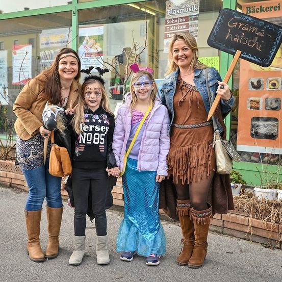 Vier Frauen und zwei Mädchen in Kostümen stehen auf einer Straße und lächeln für ein Foto. Die Frauen halten ein Schild mit der Aufschrift 'Grama haischi'. Hinter ihnen befindet sich ein Gebäude mit Glasfenstern und einem Schild, das 'Bäckerei' besagt.