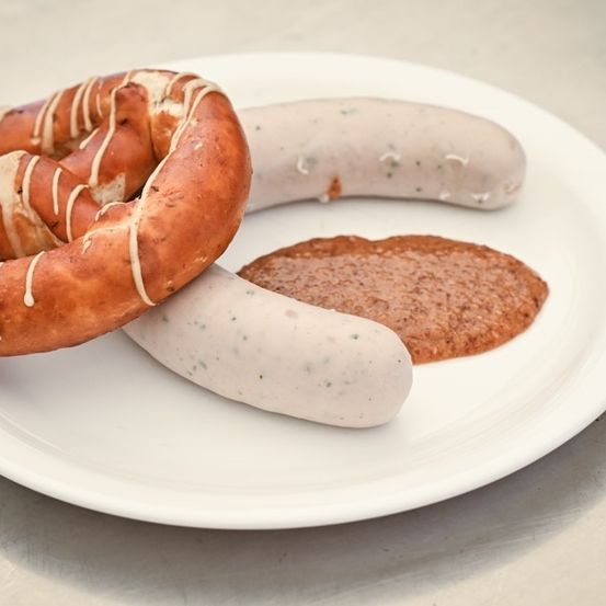 Ein weißer Teller enthält ein Brezel mit weißer Glasur, eine Wurst mit Blauschimmelkäse und eine braune Beilage.