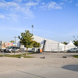Ein Parkplatz mit einem weißen Zelt, einem LKW und einem Van, die in der Nähe eines Metallzauns geparkt sind. Bäume und eine Straßenlaterne sind ebenfalls vorhanden.