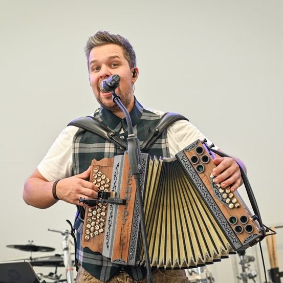 Ein Mann spielt auf der Bühne mit einem Akkordeon, trägt eine karierte Weste und hält ein Mikrofon. Hinter ihm sind ein Schlagzeug und Becken zu sehen.