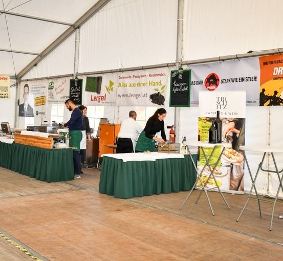 Ein Zelt-Event mit mehreren Tischen, die mit grünen Tüchern bedeckt sind. Menschen stehen hinter den Tischen, einer von ihnen hält eine Flasche. Banner hängen an den Wänden.