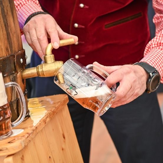 Ein Mann in traditioneller Kleidung zapft Bier aus einem Zapfhahn in ein Glaskrug auf einem Bierfest.