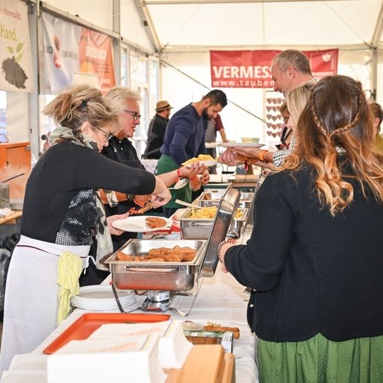 Eine Gruppe von Menschen serviert Essen an einem Buffettisch in einem Zelt. Sie tragen Schürzen und einige tragen Brillen. Es gibt mehrere Tabletts mit Essen auf dem Tisch, und einige Menschen halten Teller. Es gibt auch mehrere Tische mit Tellern und Schüsseln mit Essen. Es gibt ein Banner mit dem Text Vermes und einer Website-Adresse.