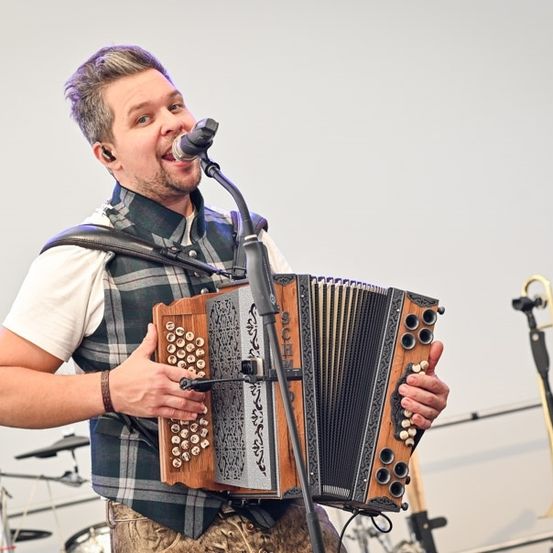 Ein Mann spielt auf der Bühne mit einem Akkordeon, singt in ein Mikrofon, trägt eine karierte Weste und ein weißes Hemd, mit einer Trompete und Trommeln im Hintergrund.