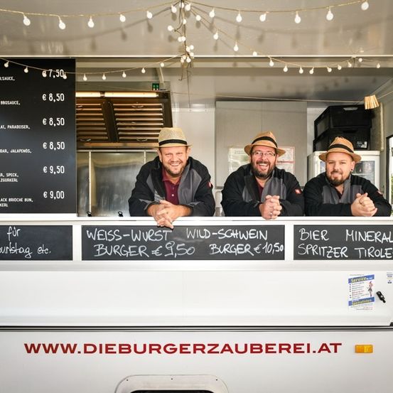 Drei Männer in Hüten stehen hinter einem Tresen eines Essenswagens mit einem Menübrett und einer Website-Adresse. Das Menü umfasst Burger, Bier und Getränke.