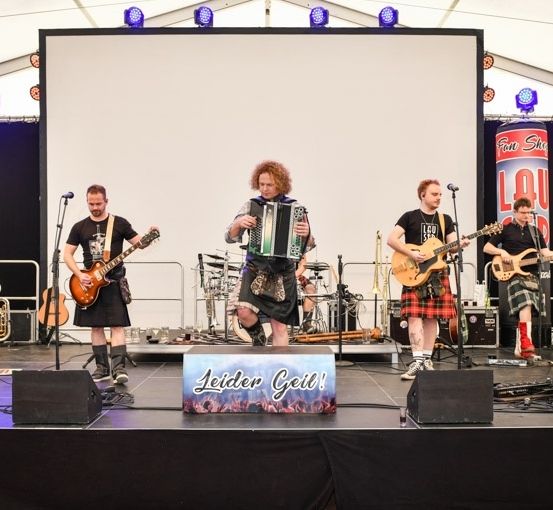Eine Band spielt auf einer Bühne mit Hintergrund. Der Leadsänger spielt Gitarre, während die anderen Akkordeon und Schlagzeug spielen. Vor ihnen steht ein Mikrofon auf einem Ständer.