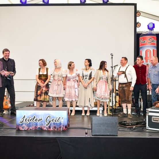 Eine Gruppe von Menschen auf der Bühne, einige in traditioneller Kleidung, mit einem Schild, das Leider Geil! und einem Hintergrund mit einem Fun Shop-Logo zeigt.