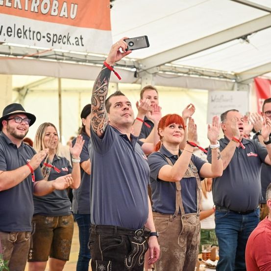 Eine Gruppe von Menschen in traditioneller bayerischer Kleidung, die unter einem Zelt mit Bannern applaudieren und lächeln, ein Mann macht ein Selfie mit einem Handy.